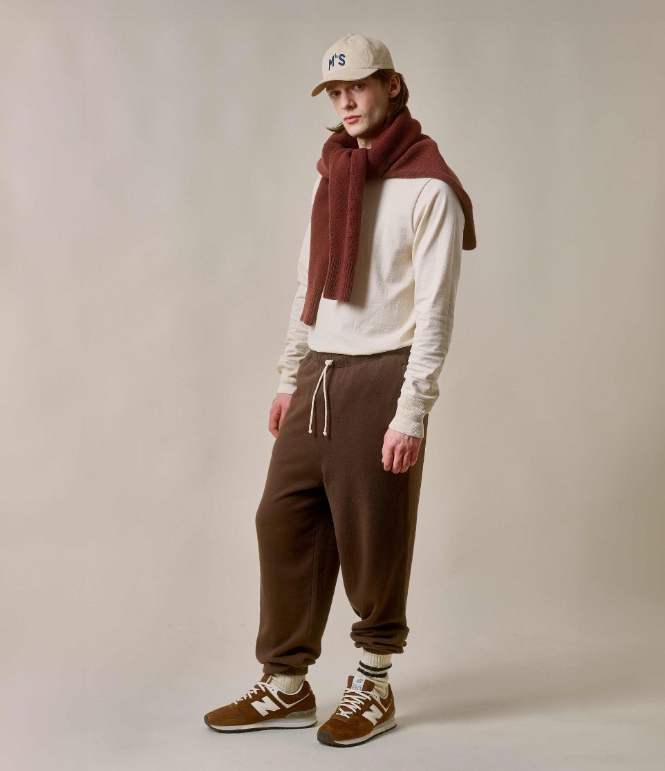 man wearing brown sweatpants, henley in nature and has knitted pullover in brown wrapped around his shoulders