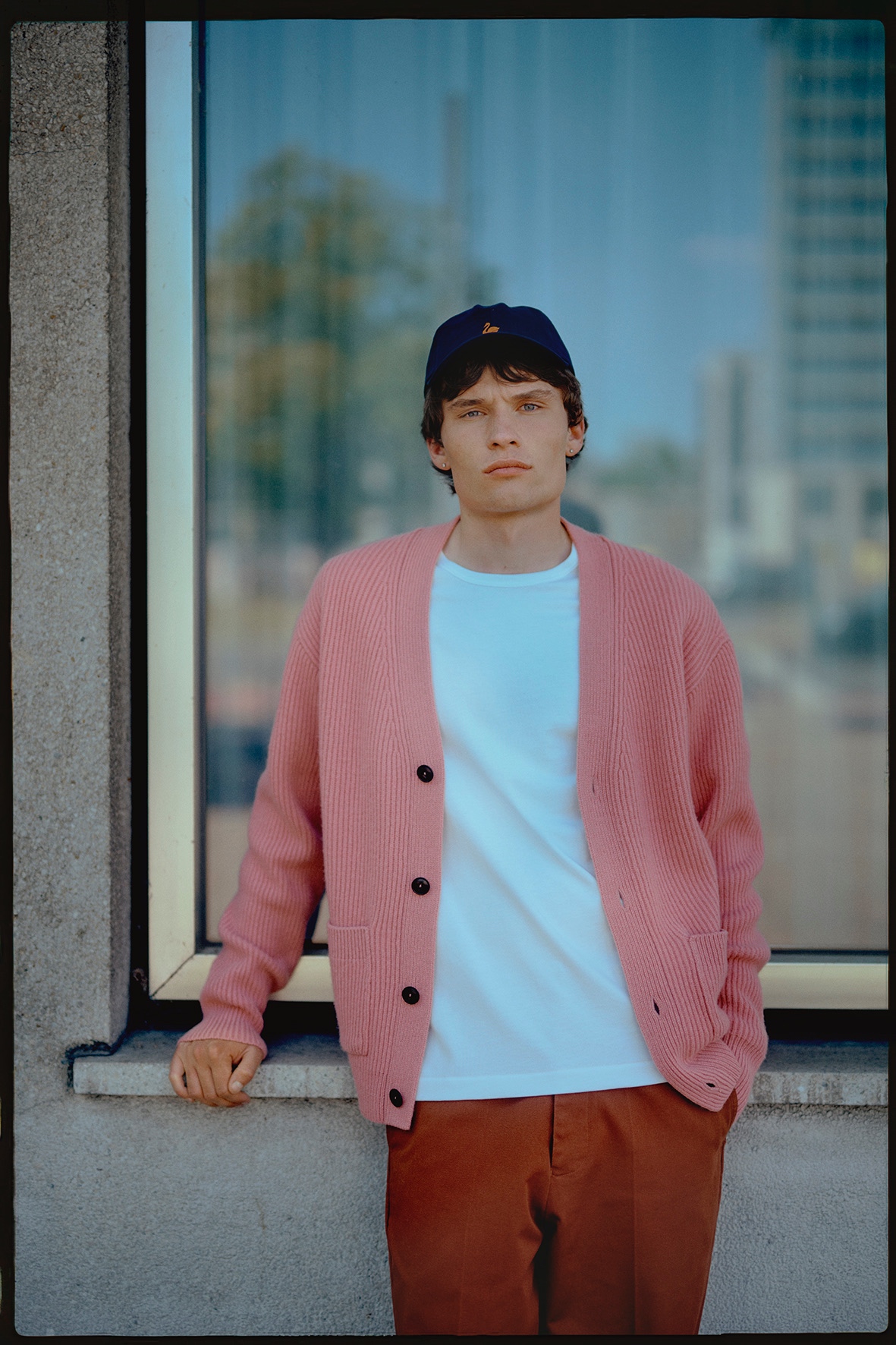 Man wearing white T-shirt, pink knit cardigan and blue cap in front of a window