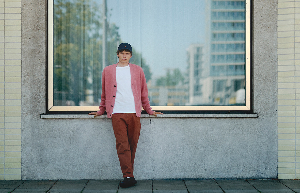 Man wearing white T-shirt, pink knit cardigan, chino pants and blue cap in front of window