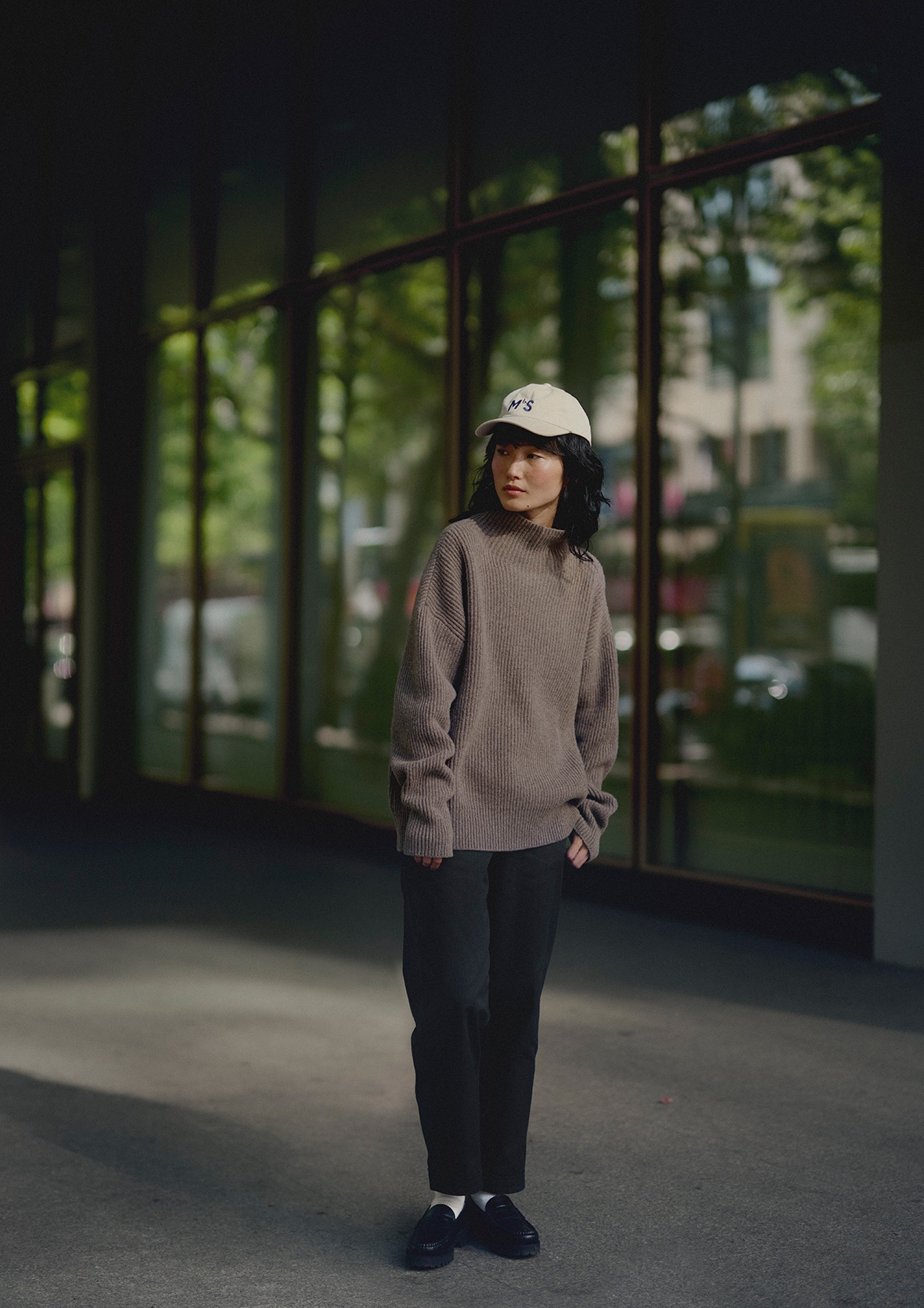 Woman wearing grey turtleneck pullover, black pants and white cap in front of a window 