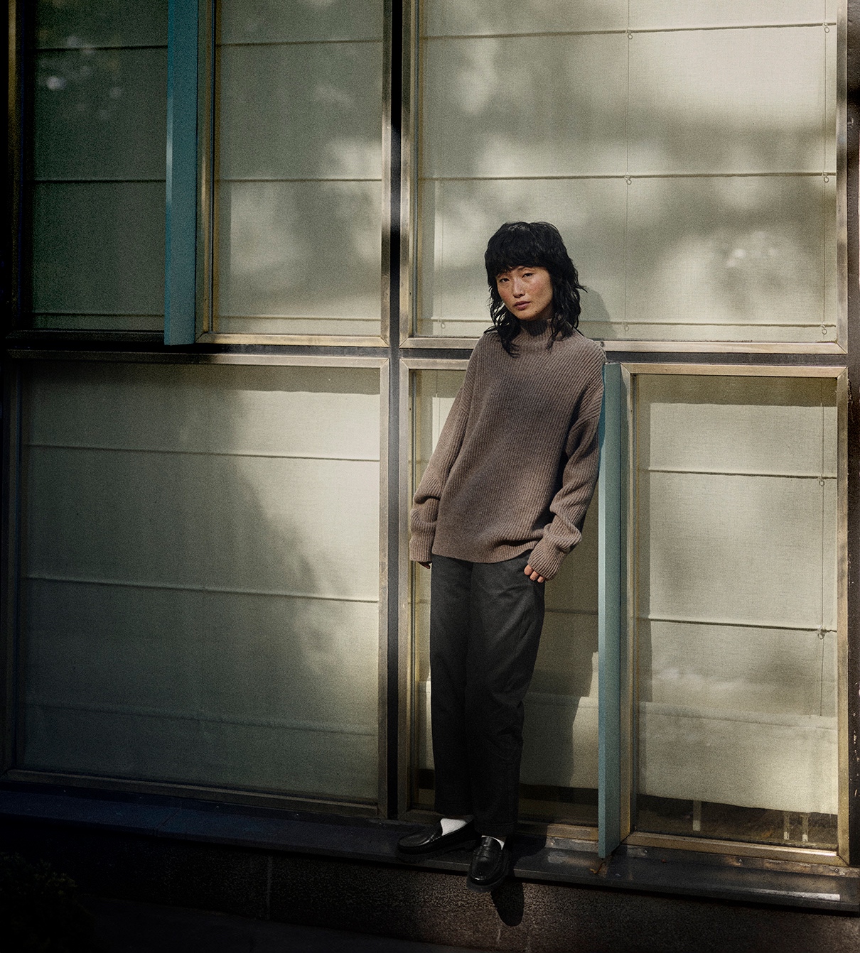 Woman wearing grey turtleneck pullover and black pants in front of a window 