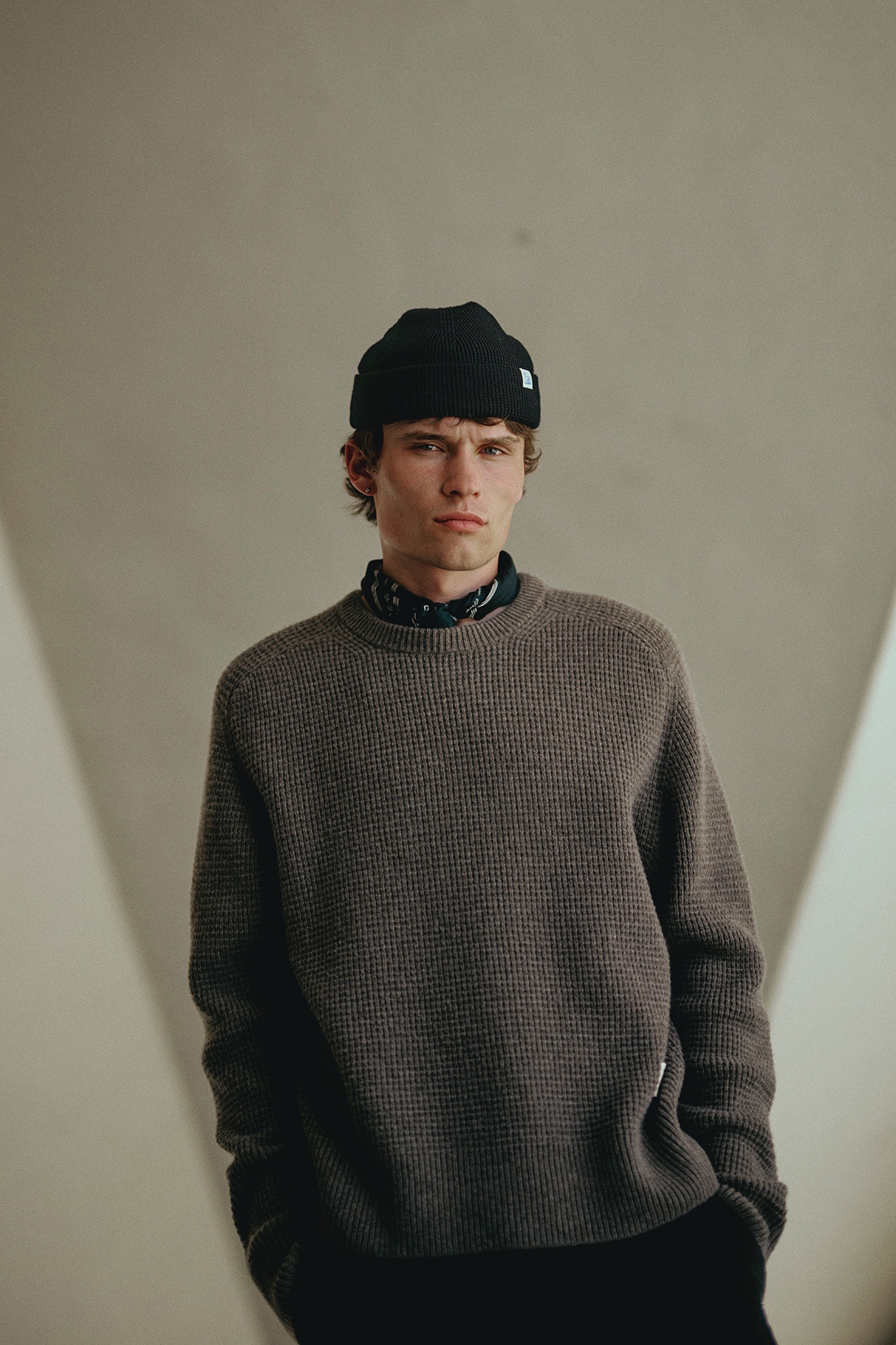 Man wearing grey crewneck pullover, black beanie and bandana in front of a wall