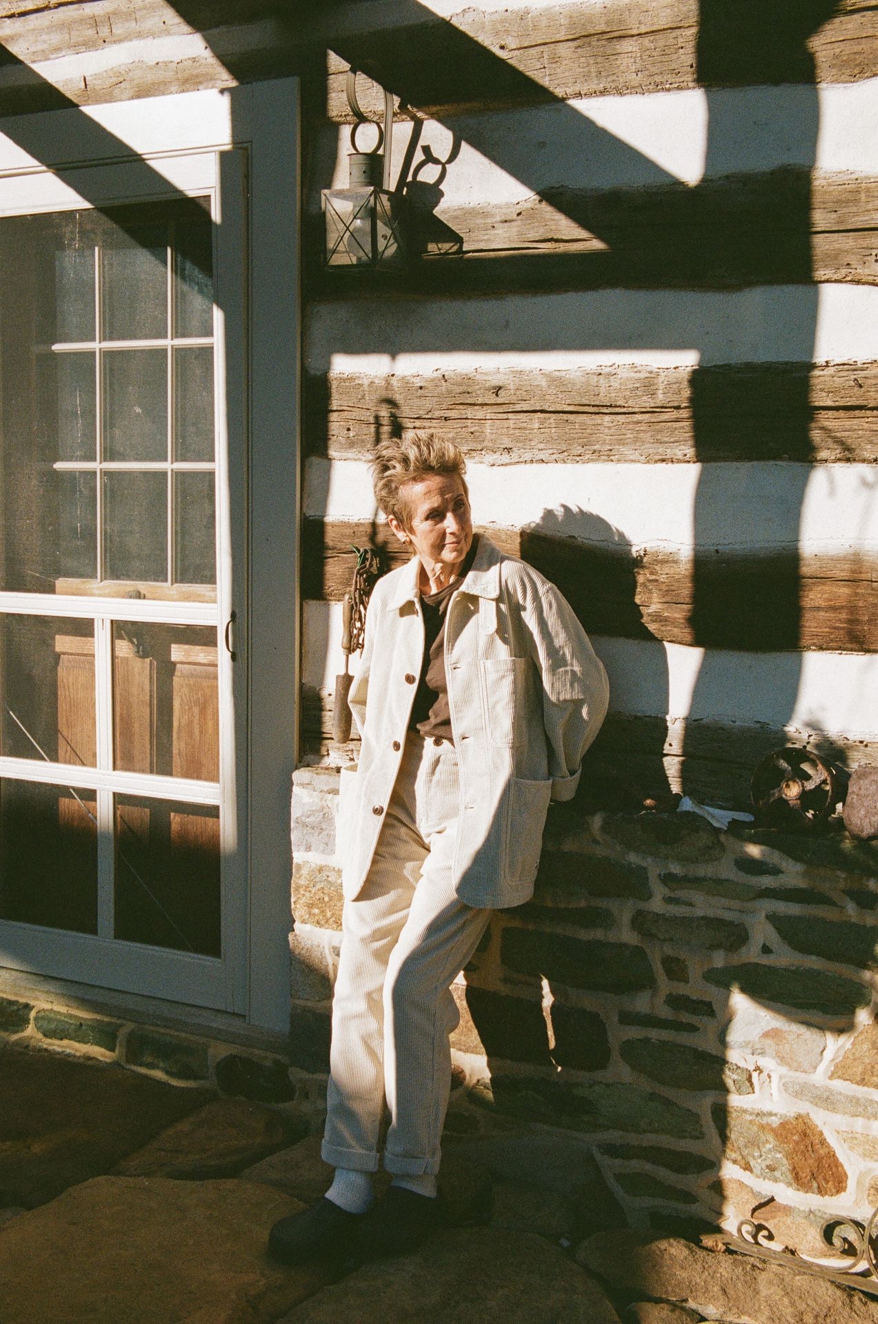 Woman in white corduroy jacket and trousers leaning on rustic wall