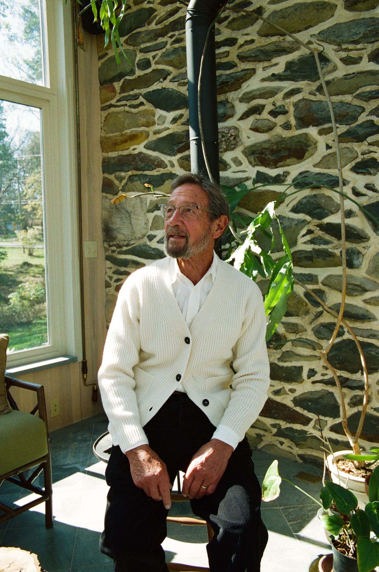 Man in white knit cardigan and shirt sitting on chair in front of stone wall